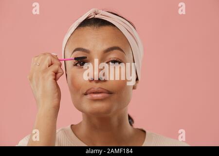 Kopf und Schultern Portrait der schönen gemischten Frau, die Mascara anwendet, während sie morgens auf Make-up setzt, vor staubrosa Hintergrund stehend, kopiere Raum Stockfoto