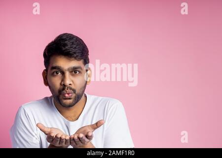 Gutaussehender indischer männlicher Blasluftkuss, der seine Liebe, seine Sympathie für die Freundin zum Ausdruck bringt, mit einem beiläufigen weißen T-Shirt, isoliert über dem rosafarbenen Hintergrund mit c Stockfoto