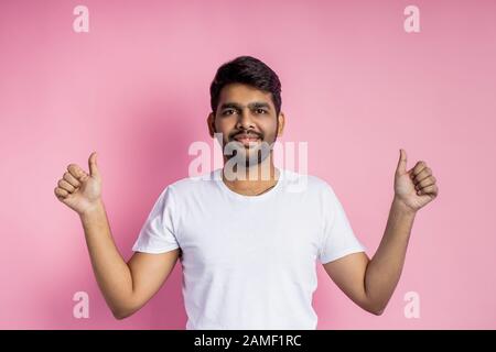 Zufriedener junger indischer Mann mit weißem T-Shirt, mit Daumen nach oben oder wie Geste, seine Zustimmung zeigend, mit beiden Händen gestikulierend, lächelnd aussehend Stockfoto