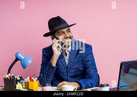 Fröhlicher stilvoller indischer Unternehmer, der formelle Kleidung trägt, schwarzen Hut, auf dem Handy spricht, breit lächelt, nach Seite schaut, am Schreibtisch im Offic sitzt Stockfoto