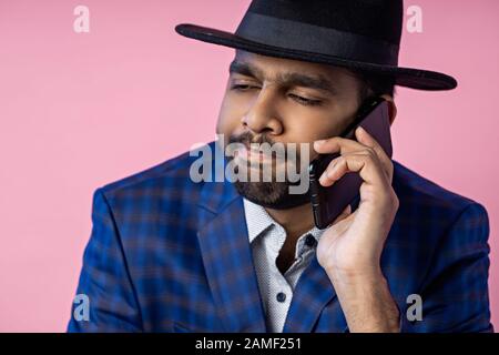 Besorgter junger indischer bärtiger Geschäftsmann, der auf dem Handy spricht. Nahaufnahme des Mannes in formeller Kleidung, schwarzer Hut, mit ernsthaftem, konzentriertem Aussehen während ha Stockfoto
