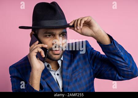 Nahaufnahme des eleganten indischen Unternehmers, der blau karierte Anzug trägt, den Hut berührt, auf dem Handy mit ernsthaftem Ausdruck spricht, während er sitzt Stockfoto