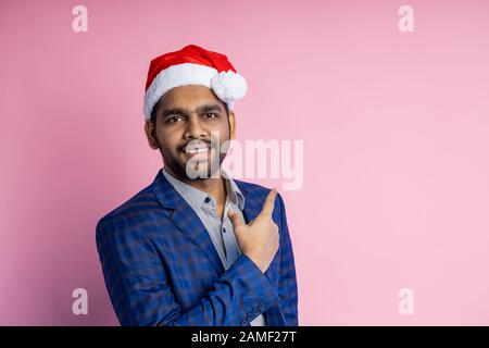 Horizontaler Schuss des gefielen indianischen Unshaven, mit Brille, blau kariertem Anzug, Weihnachtsmann, lächelnd mit der Kamera blickend, mit Zeigefinger b Stockfoto