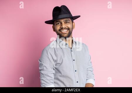 Taille hoch Portrait von fröhlichen fröhlichen, gutaussehenden jungen unrasierten indianern mit Hemd, modischem schwarzen Hut, mit breitem Lächeln und Standin auf die Kamera blicken Stockfoto