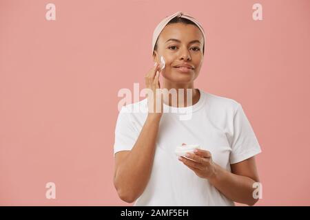 Taille hoch Portrait von Mixed-Race junge Frau mit Gesichtscreme und Lächeln auf der Kamera, während sie sich morgens vor pinkfarbenem Hintergrund während der Schönheitsroutine posiert Stockfoto