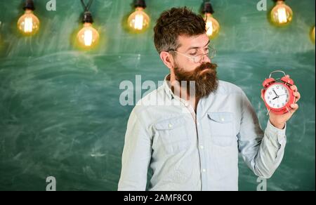 Schulzeitplan Konzept. Mann mit Bart und Schnurrbart auf interessiertes Gesicht überprüft die Zeit. Bärtiger Hüpfer hält Uhr, Schwarzen Brett im Hintergrund, Kopierraum. Lehrer in Brillen hält Wecker. Stockfoto