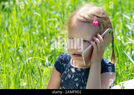 Süßes kleines Mädchen spricht am Telefon. Ein kleines Mädchen sitzt mit einem Handy an einem sonnigen Sommertag auf einer grünen Wiese. Das ernste Gesicht eines Kindes Stockfoto