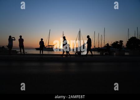 Silhouetten von Menschen, die den Sonnenaufgang und die Yacht-Masten bewundern, die von goldenem Stundenlicht in Naxos Town, Cyclades Group of Islands Greece, beleuchtet werden. Stockfoto