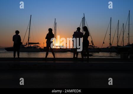 Silhouetten von Menschen, die den Sonnenaufgang und die Yacht-Masten bewundern, die von goldenem Stundenlicht in Naxos Town, Cyclades Group of Islands Greece, beleuchtet werden. Stockfoto