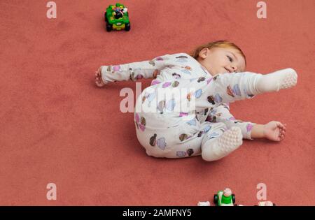 Glückliches achtzehnmonatiges altes Baby auf dem Rücken, das versucht, auf den Boden zu rollen. Frühe Entwicklung von Kindern. Stockfoto