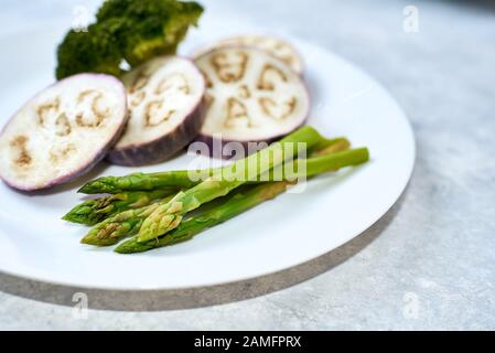 Gedünstetes Gemüse in einer weißen Platte auf einer blauen Tabelle. Auberginen, Brokkoli, Spargel. Stockfoto