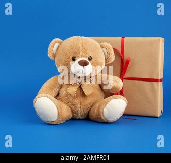 Beigefarbener Teddybär und eine Schachtel in braunem Kraftpapier verpackt und mit rotem Band, festlicher Kulisse für Valentinstag und Geburtstag verbunden Stockfoto