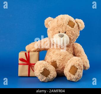 Der große beige Teddybär hält eine Schachtel in braunem Papier und mit rotem Band, festlicher Kulisse für Valentinstag und Geburtstag. Saisonale Geschäftstage Stockfoto