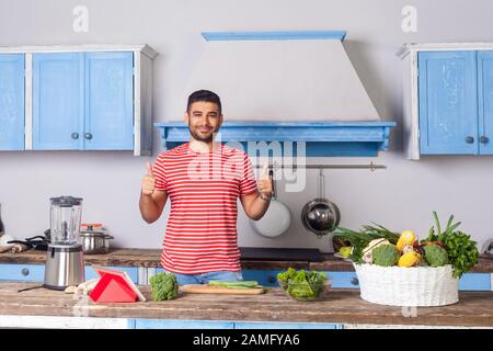 Positiver Mann mit Daumen nach oben wie Geste in der modernen Küche, lächelnde Kamera, zufrieden mit vegetarischer Kost, veganer Ernährung, Korb mit frischem Grün Stockfoto