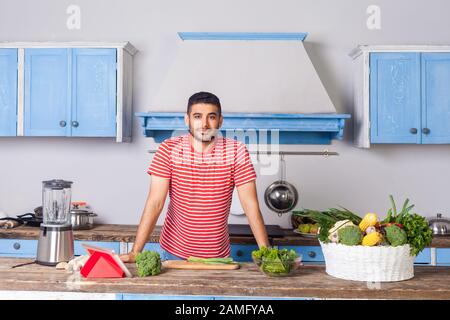 Junger gutaussehender Mann, der in einer Küche mit modernen Möbeln steht und mit Lächeln auf die Kamera blickt, Korb mit frischem grünen Gemüse und Mixer auf dem Tisch, VEG Stockfoto