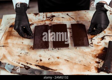 3/4-Blick auf Chocolatier in Latexhandschuhen in der Nähe von Schokoladenriegeln Stockfoto