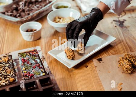 3/4-Blick auf Chocolatier in schwarzem Latexhandschuh, der leckere Süßigkeiten auf den Teller legt Stockfoto