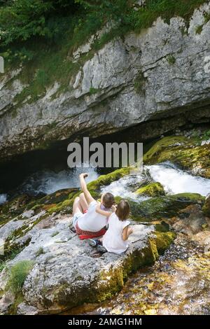 Mutter mit Kindern, die ein selfie nehmen, umarmen und auf einem Felsen an einem Bergbach auf einem Familienausflug sitzen. Outdoor-Lifestyle, natürliche Erziehung, Kindheit Stockfoto