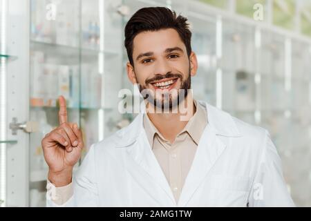 Gutaussehender Apotheker lächelt an der Kamera und zeigt mit dem Finger in der Apotheke Stockfoto