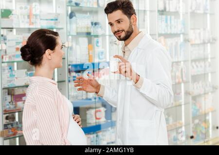 Lächelnder Apotheker, der schwangeren Frau einen Mixbecher mit Pillen von Drogerien zeigt Stockfoto