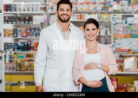 Lächelnder Apotheker und schwangere Frau, die im Hintergrund Kamera mit Apothekenvorschau betrachtet Stockfoto