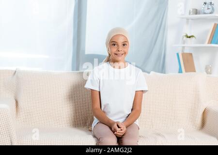 Glückliches Kind im Kopftuch, das auf dem Sofa im Wohnzimmer sitzt Stockfoto