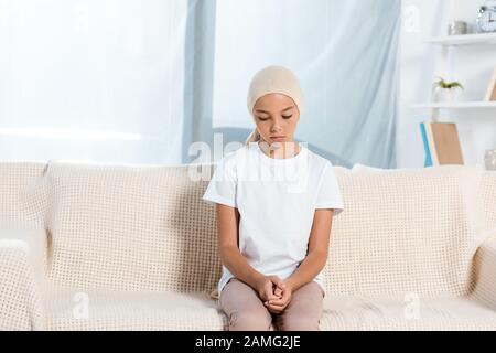 Verärgertes Kind im Kopftuch, das auf dem Sofa im Wohnzimmer sitzt Stockfoto