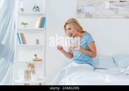 Schockierte Frau, die digitales Thermometer hält, während sie zu Hause auf dem Bett sitzt Stockfoto