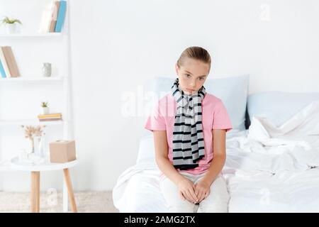 Krankes Kind im Schal, das zu Hause auf dem Bett sitzt Stockfoto