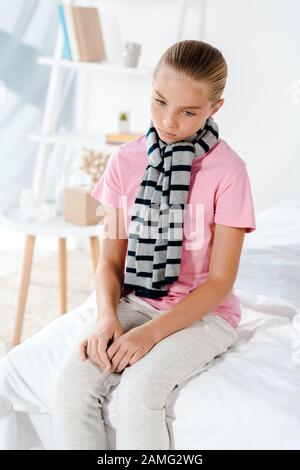 Krankes Kind im gestreiften Schal, das zu Hause auf dem Bett sitzt Stockfoto