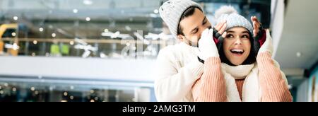 Panoramaaufnahme des fröhlichen Freundes, der die Augen vor aufgeregter Freundin schließt, um auf der Eislaufbahn eine Überraschung zu machen Stockfoto