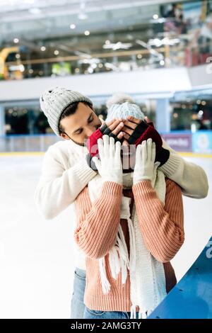 Fröhlicher Mann, der die Augen vor der Frau schließt, um auf der Eisbahn eine Überraschung zu machen Stockfoto