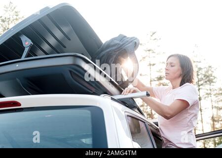 Ernsthafte, fokussierte Frau legt Dinge in den Kofferraum auf das Dach eines Autos oder in einen Frachtkasten. Vorbereitung für einen Familienurlaub. Stockfoto