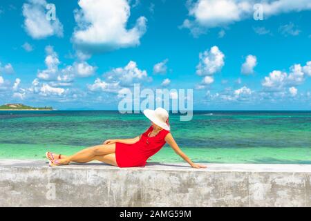 Luxus-Tourismus auf den Seychellen. Elegante Lifestyle-Frau in rotem Kleid, die an einer niedrigen Wand mit türkisfarbenem Wasser im Indischen Ozean des schönen Anse Kerlan liegt Stockfoto