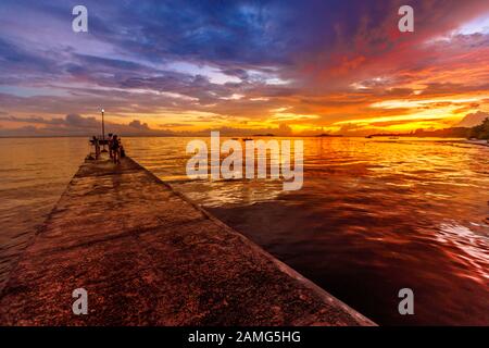 Holzsteg mit Sonnenuntergang, der sich im tropischen Meer widerspiegelt. Grand Anse Fond de Lanse Strand am Indischen Ozean auf der Insel Praslin, Seychellen. Stockfoto