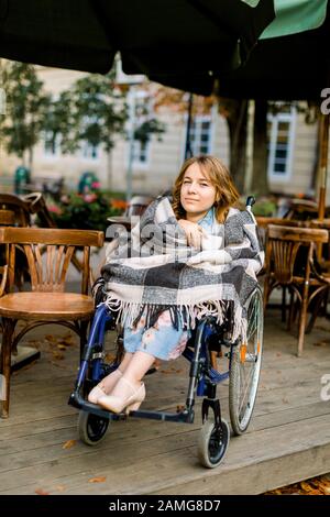 Portrait einer jungen, schönen Frau im Rollstuhl, bedeckt mit karierter Pflauge, Kaffee trinken, während sie im Café draußen sitzt. Stockfoto