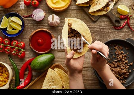 3/4-Ansicht des Mannes, der Hackfleisch in Taco mit rohen Zutaten auf Holzoberfläche hinzufügt Stockfoto