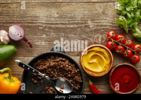 Draufsicht auf Hackfleisch in Bratpfanne mit Soßen und rohem Gemüse auf Holzgrund Stockfoto