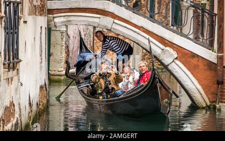 Eine Gruppe von Touristen in einer Gondel, die unter einer kleinen Brücke vorbeiführt und fast alle darauf absehen, Fotos zu machen, anstatt das Erlebnis zu genießen. Stockfoto