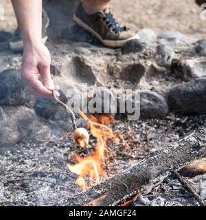 Schmackhaftes, süßes Marshmallow auf einer krummen Perücke wird von einer männlichen Hand über einem Lagerfeuer gehalten. Dessert am Lagerfeuer kochen. Stockfoto