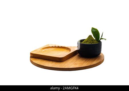 Isolieren Sie Heap Green Teepulver in einer schwarzen Keramikschale. Nehmen Sie Teeblätter in einer Tasse aus dunklem Stein heraus. Mit Mini-grünen Teeblättern auf leerer Holzplatte. Stockfoto