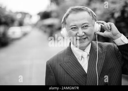 Porträt eines reifen asiatischen Geschäftsmannes, der Anzug trägt, während er im Freien Musik auf den Straßen hört Stockfoto