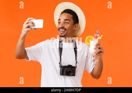 Fröhlicher, charismatischer, gutaussehender afroamerikanischer Mann in Hut, Kamera und weißem T-Shirt, der einen Glascocktail aufwirft und einen erfrischenden Drink in der Nähe der Küste genießt Stockfoto