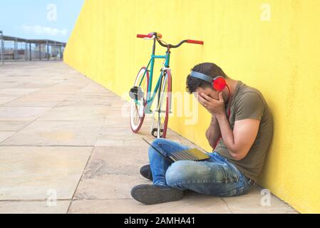 Der junge Mann verlor sich in Depressionen, die auf der Straße auf dem Boden saßen, und litt unter emotionalen Schmerzen, Traurigkeit und einem zerstörten und verzweifelten Anlehnen an die Wand Stockfoto