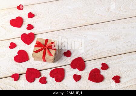 Geschenkschachtel aus Kunsthandwerk mit roter Schleife und Streuung der valentinsherzen auf einer weißen, strukturierten Holzoberfläche. Grußkarte Valentine mit Platz zum Kopieren. Stockfoto