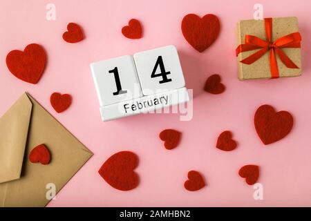 Weißer Kalender für unbefristete Tage mit Datum 14. februar, Briefumschlag aus Kunsthandwerk, Geschenkbox mit rotem Band und valentinsherzen auf einer rosafarbenen Oberfläche. Grußkarte. Stockfoto