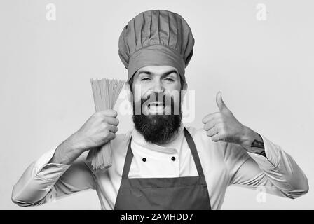 Der Küchenchef hält einen Haufen Spaghetti in der Hand. Catering und italienisches Speisekonzept. Mann mit Bart isoliert auf weißem Hintergrund. Mit gesundem Gesicht in Burgunderuniform kochen, hält trockene Pasta, die Daumen nach oben zeigen Stockfoto