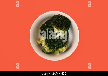 Eine weiße Schüssel mit gedämpftem grünem Brokkoli in der Mitte auf einem orangefarbenen Karotten-Farbhintergrund, Draufsicht auf ein Gericht mit gekochtem vegetarischem Gemüse Stockfoto