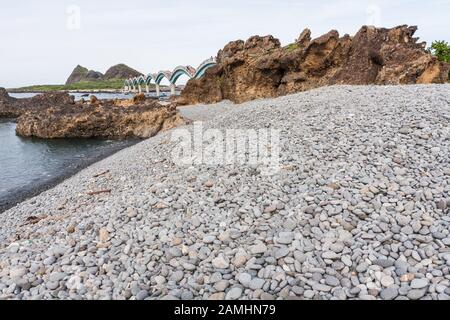 Blick auf einzigartige Felsformationen, Kieselsteine am Strand, die berühmte Fußgängerbrücke mit acht Bögen und eine kleine Insel vor der Küste in Sanxiantai in Taitung, Taiwan Stockfoto