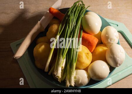 Zutaten für den Salat - gekochte Eier, Kartoffeln, Karotten und grüne Zwiebeln auf einem blauen Teller vor dem Schneiden Stockfoto